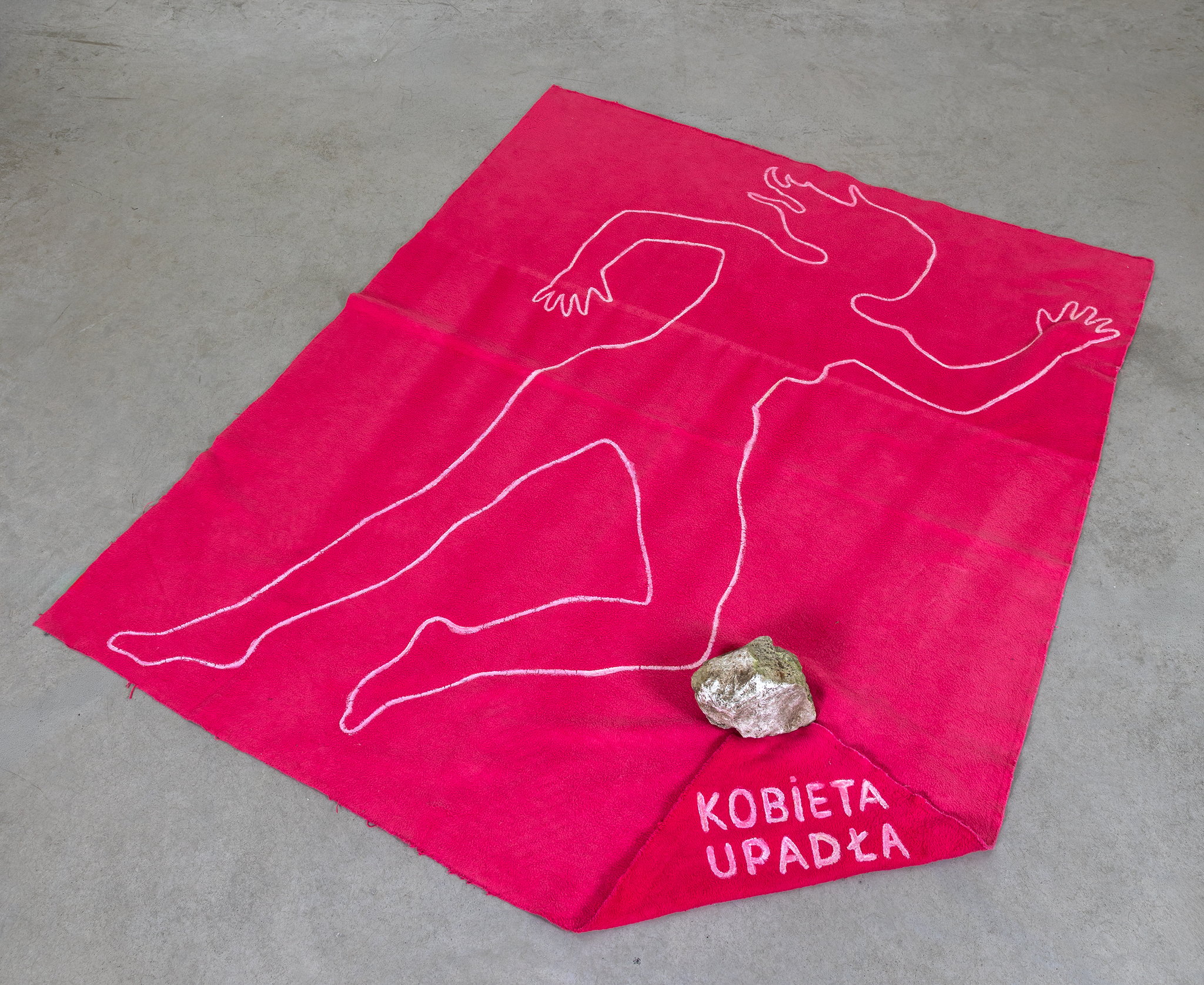 Różowa tkanina z obrysem kobiecej sylwetki, kamieniem i napisem Kobieta upadła