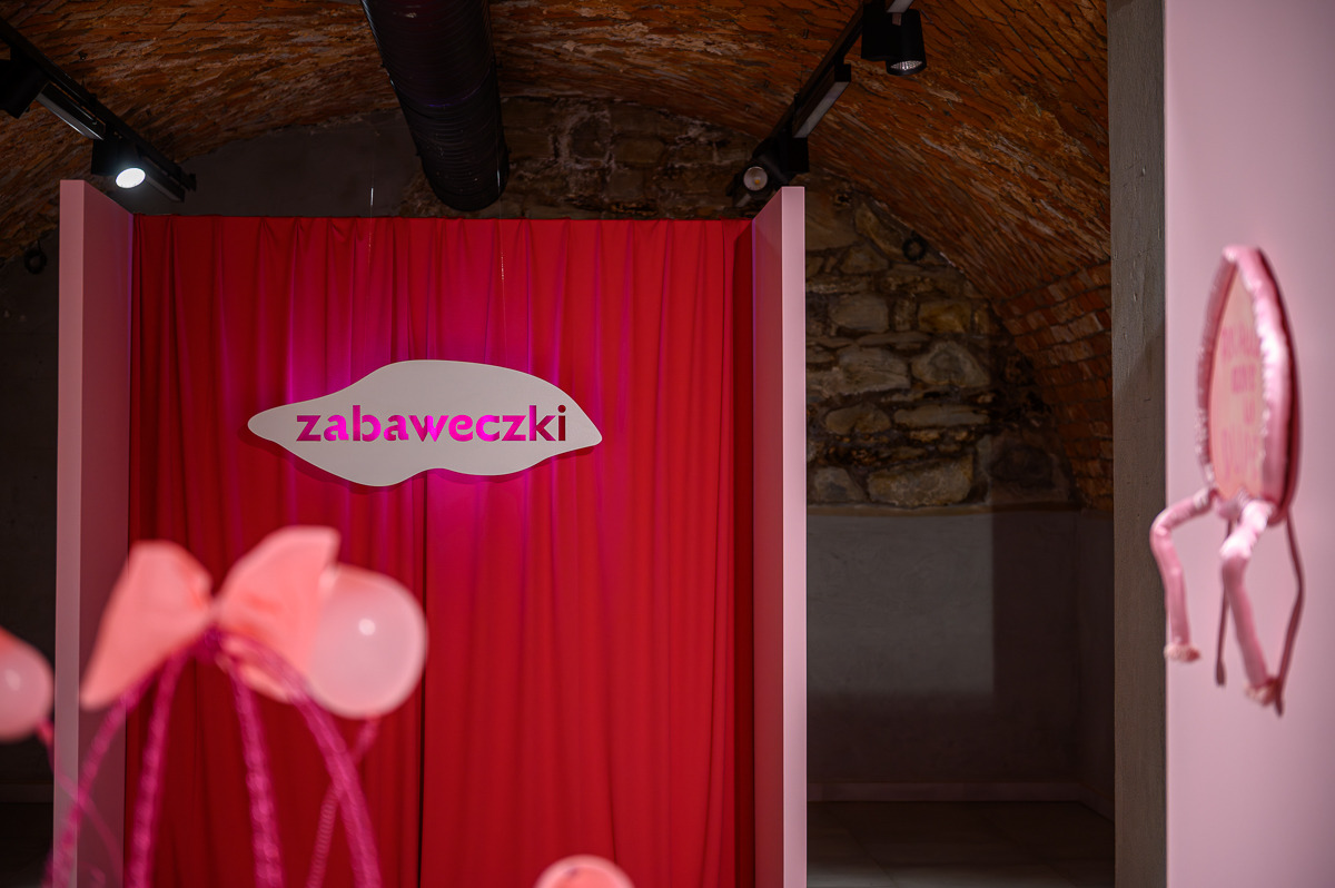 Czerwona kurtyna z&nbsp;napisem „zabaweczki” i&nbsp;różowe obiekty na&nbsp;wystawie Kamili Jajkowskiej
