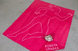 Różowa tkanina z obrysem kobiecej sylwetki, kamieniem i napisem Kobieta upadła