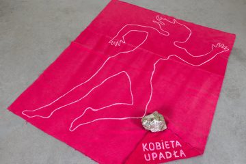 Różowa tkanina z obrysem kobiecej sylwetki, kamieniem i napisem Kobieta upadła