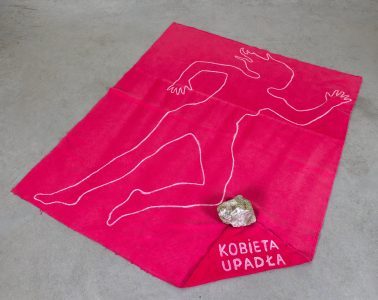 Różowa tkanina z obrysem kobiecej sylwetki, kamieniem i napisem Kobieta upadła